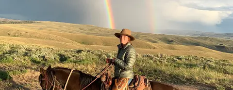 A person on horseback smiling with a double rainbow in the sky over rolling hills.