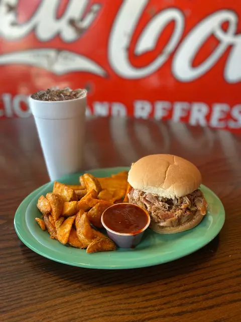 A plate of pulled pork sandwich, potato wedges, and a drink on a wooden table.