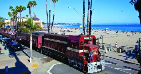 A red train travels along a street beside a beach.