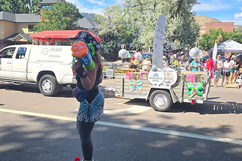 A person in a street parade holds a toy water gun near a decorated trailer.