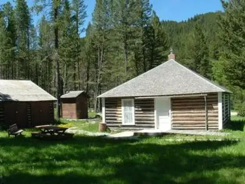 Wooden cabins stand in a grassy clearing surrounded by tall trees.