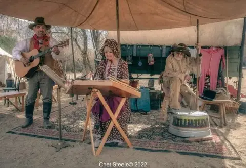 Three people in historical clothing play musical instruments under a tent outdoors.