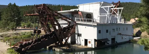 A large historic dredge sits in a body of water near a wooded area.