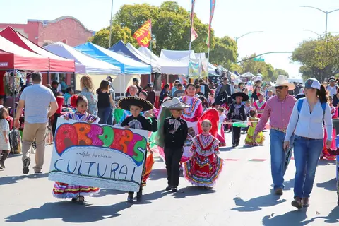 Children and adults in traditional clothing walk in a street parade past vendor booths.