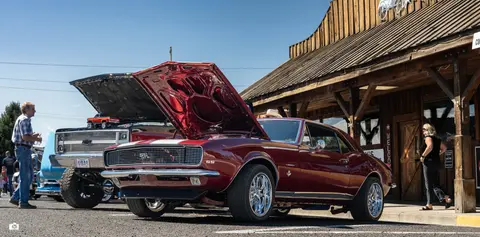 A classic red car with its hood open is parked outside a rustic wooden building.