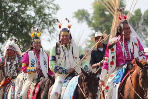 People in traditional regalia ride horses outdoors.