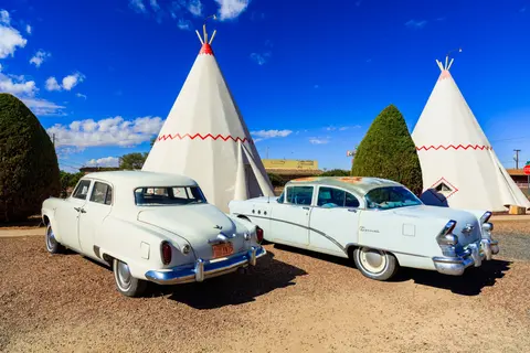 Classic cars parked in front of teepees under a blue sky.