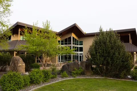 Exterior view of a modern building with large glass windows, surrounded by landscaping, trees, and boulders.