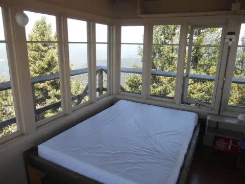 Interior view of a lookout tower with a bed surrounded by windows overlooking trees.