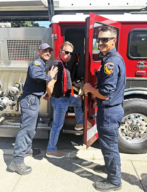 Two firefighters playfully posing with a man by a fire truck.
