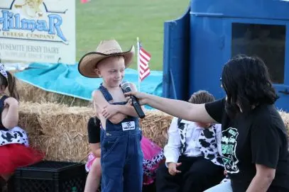 A young boy in a cowboy hat and overalls speaks into a microphone held by a woman.
