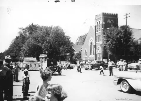 Black and white historic photo showing people and vintage cars on a street beside a church.