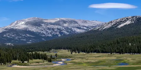 A scenic view of a wide valley with a winding river, dense forest, and mountains under a blue sky with a single lenticular cloud.