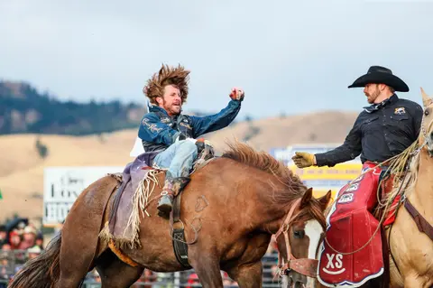 A cowboy rides a bucking horse while another rider looks on.