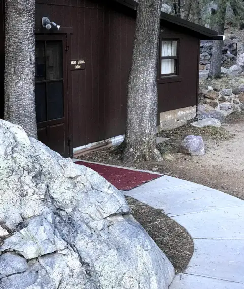 Exterior view of a dark brown cabin with trees and a rocky path in front.