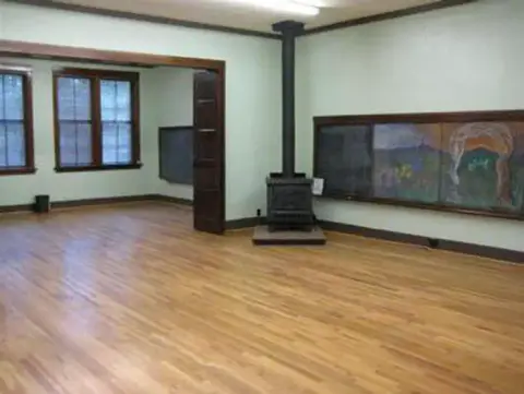 An interior view of a historic schoolroom with wooden floors and a wood stove.