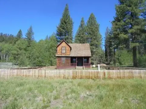 A wooden house stands surrounded by a wooden picket fence among trees and grass.
