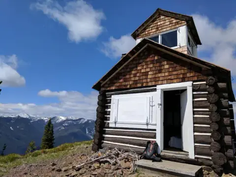 A rustic log cabin stands on a mountain ridge under a blue sky.