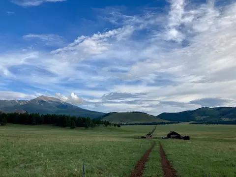 A dirt track leads across a green meadow toward a cabin beneath a mountain and a cloudy sky.