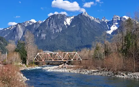 A rusty metal bridge crosses a river with snow-capped mountains in the background.