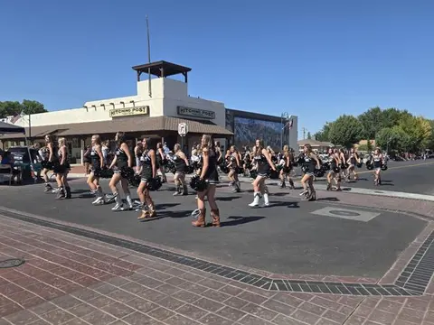 A group of performers in black outfits and boots march down a street in front of a building.