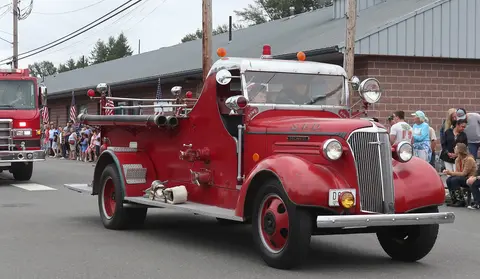 A vintage red fire truck drives down a street during an outdoor event.