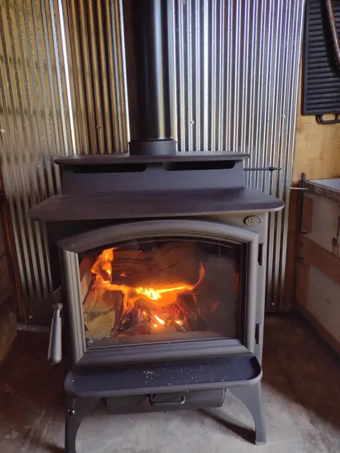 A wood-burning stove is lit indoors against a corrugated metal wall.