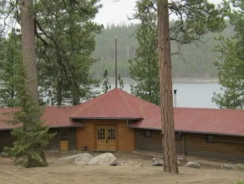 A single-story wooden building with a red roof stands among pine trees near a body of water.