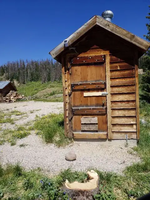 A small wooden outhouse stands outdoors under a clear blue sky.