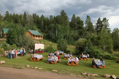 People enjoying an outdoor picnic and meal on the lawn near wooden cabins and a covered wagon.