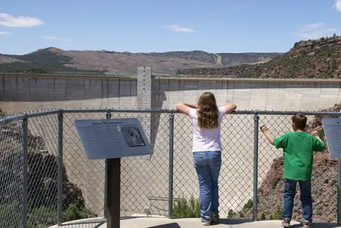 Two children look at a large concrete dam from behind a chain-link fence.