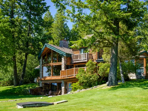 Exterior view of a large log cabin surrounded by trees and a green lawn.
