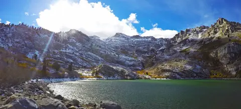 A scenic view of a lake surrounded by snow-dusted mountains under a bright cloudy sky.