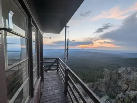 A view from the balcony of a fire lookout tower overlooking a forest and mountains at sunset.