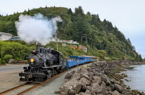 A steam locomotive pulls passenger cars along a coastal railway.