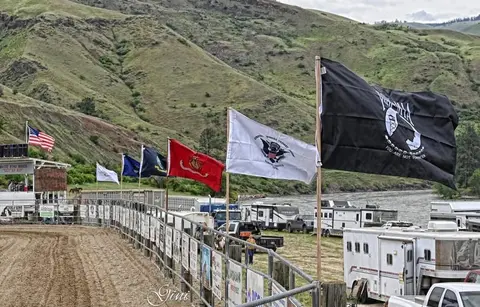 A line of flags stands along a fenced arena with rolling green hills and a river in the background.