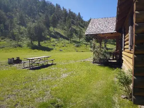 A log cabin sits next to a grassy lawn with a picnic table and a forested hill in the background.