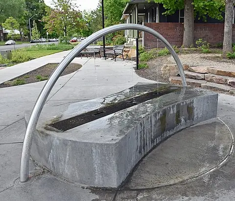 A modern arched water fountain sits next to a sidewalk and a building.