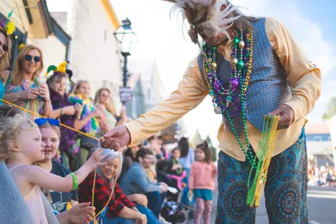 A person in a feathered mask and bead necklaces hands beads to a child in a crowd.