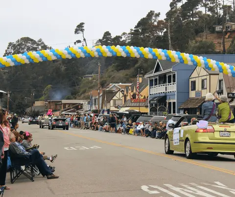 A parade with cars and crowds along a street decorated with a balloon arch.