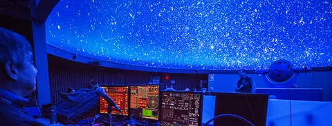 A view of the interior of a planetarium with a domed starry ceiling and control panels in the foreground.