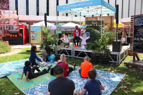 People sit on mats on the lawn watching a speaker on a stage.