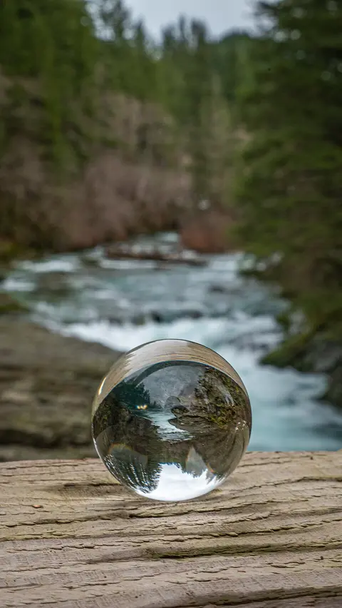 A crystal ball rests on a wooden surface with a blurred river and forest in the background.
