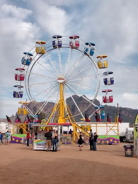 A colorful Ferris wheel stands outdoors at a fair with mountains in the background.