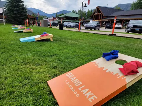 Cornhole boards set up on a grassy area in front of buildings and mountains.