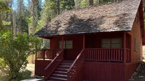 A red wooden cabin with a shingled roof stands surrounded by trees.
