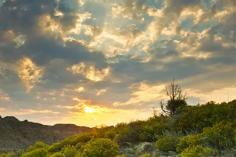 A sunset over a hilly landscape with sparse vegetation and a dramatic cloudy sky.