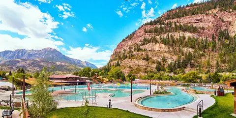 A hot springs resort pool area sits below large rocky mountains under a bright blue sky.