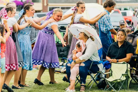 A group of dancers in matching dresses perform outdoors near seated spectators.