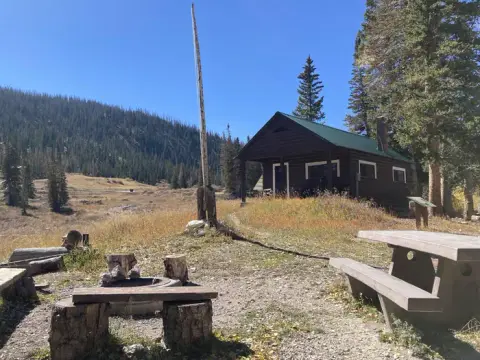 A rustic log cabin stands in a mountain landscape with a picnic table and fire ring in the foreground.